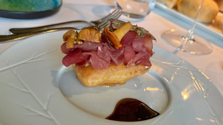 A gourmet dish featuring a fried base topped with thin slices of cured meat, garnished with mushrooms and herbs, served on a white plate with a dark sauce on the side.