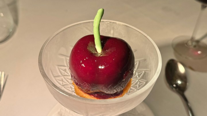 A dessert resembling a shiny red apple with a green stem, set on a delicate transparent plate.