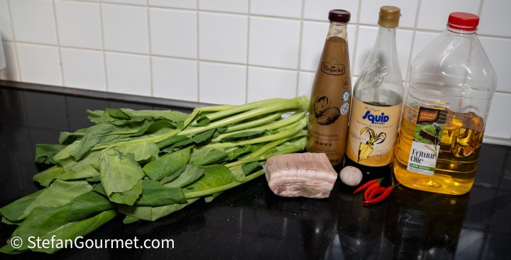 A selection of fresh spinach leaves, a piece of pork belly, garlic, red chilies, and various cooking oils and vinegars arranged on a black countertop.
