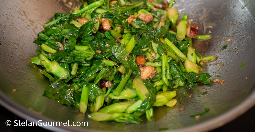 A close-up view of sautéed greens, including leafy vegetables and chopped pieces, mixed in a silver frying pan.