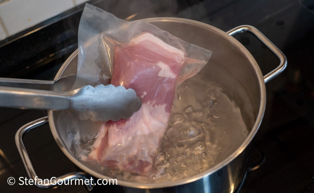 A vacuum-sealed bag containing meat is being submerged in a pot of boiling water, with tongs holding the bag.