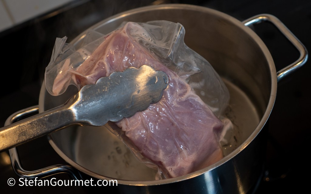 A piece of meat in a vacuum-sealed bag is being cooked in a pot of steaming water, held by tongs.