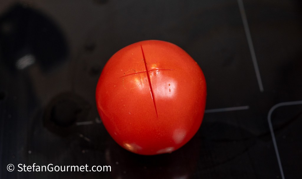 A whole red tomato with a cross incision on its surface, placed on a dark countertop.