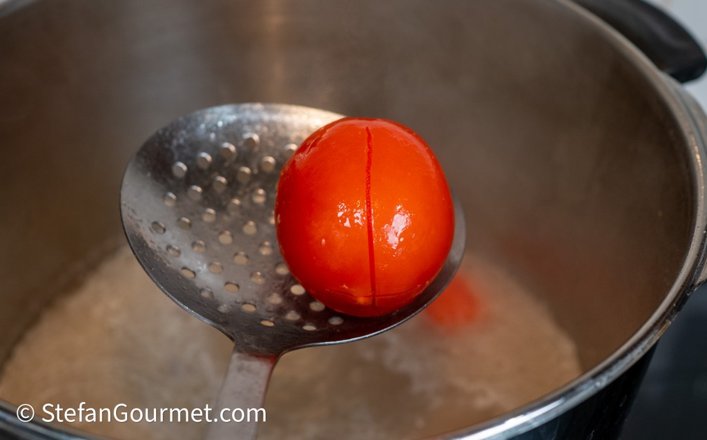 A whole ripe tomato being lifted from a pot of boiling water using a slotted spoon.