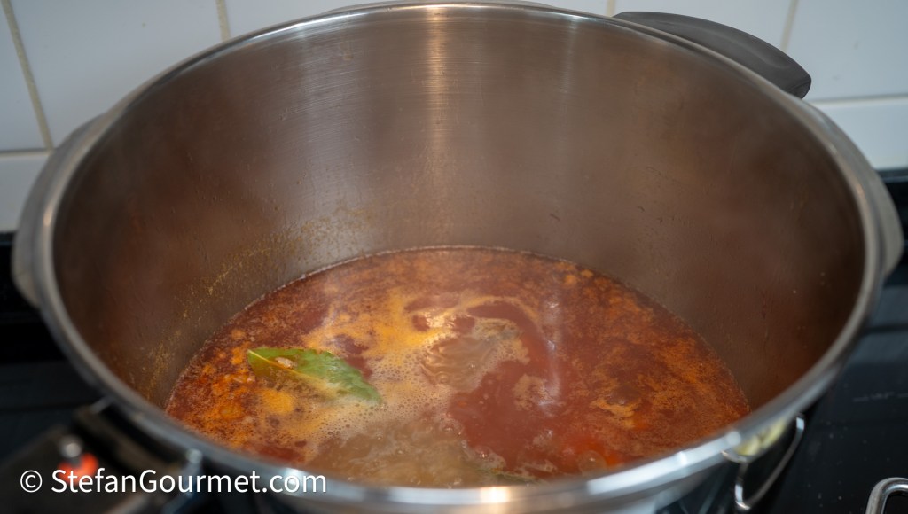 A pot filled with simmering broth, featuring bits of spice and a bay leaf floating on the surface.