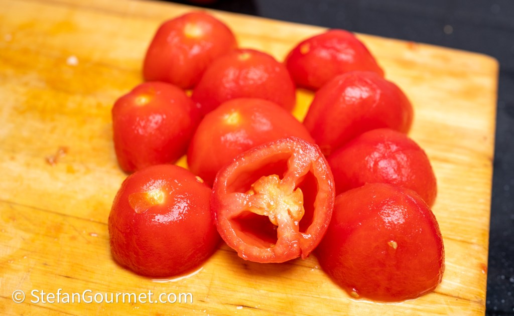 A wooden cutting board with several peeled tomatoes, some whole and one cut in half, showcasing the seeds inside.