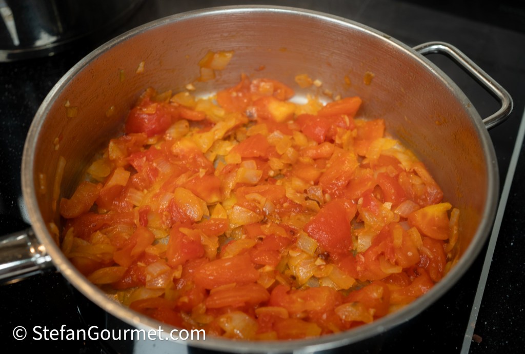 A pot containing sautéed tomatoes and onions, with a mix of colorful diced bell peppers, cooking on a stovetop.