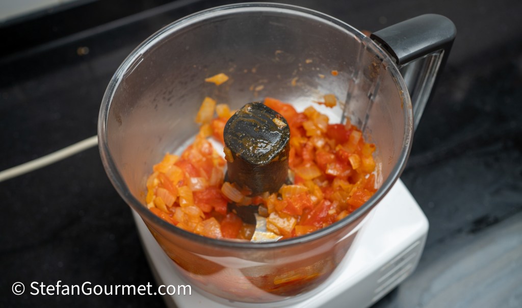 A food processor containing chopped tomatoes and bell peppers on a black countertop.