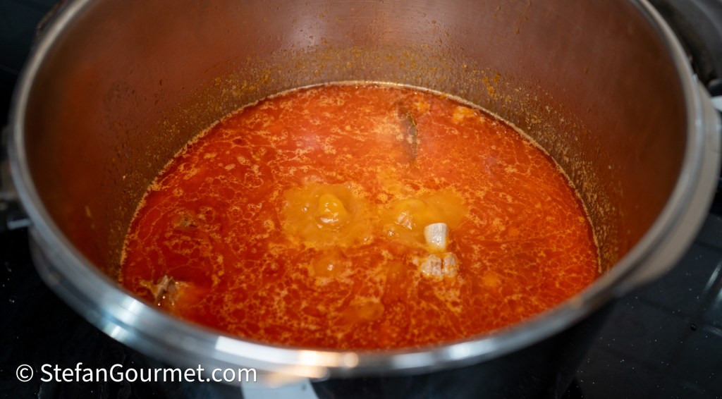 Close-up view of simmering stew in a stainless steel pot with a rich red broth and floating ingredients.