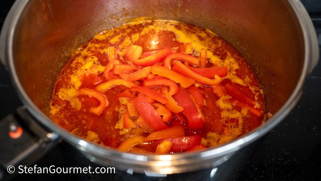A close-up of a pot containing simmering red and yellow bell peppers in a thick, spicy sauce.
