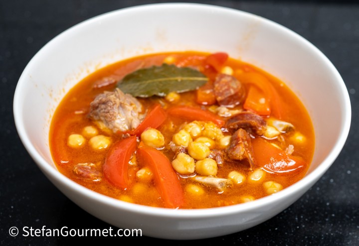 A bowl of hearty stew containing chickpeas, red bell peppers, pieces of meat, and herbs in a rich tomato-based broth.
