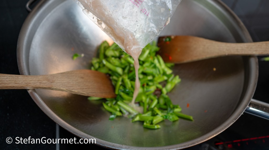 Pouring liquid from a plastic bag into a skillet with chopped green peppers and wooden spatulas.