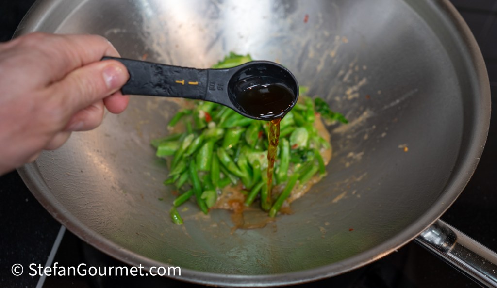 A hand holding a measuring spoon pouring liquid into a wok with green vegetables and sauce.