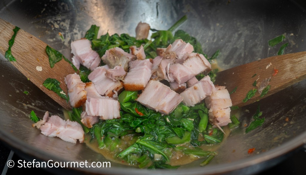 A close-up of a wok filled with sautéed green leafy vegetables and chunks of cooked pork, with wooden spatulas blended in.