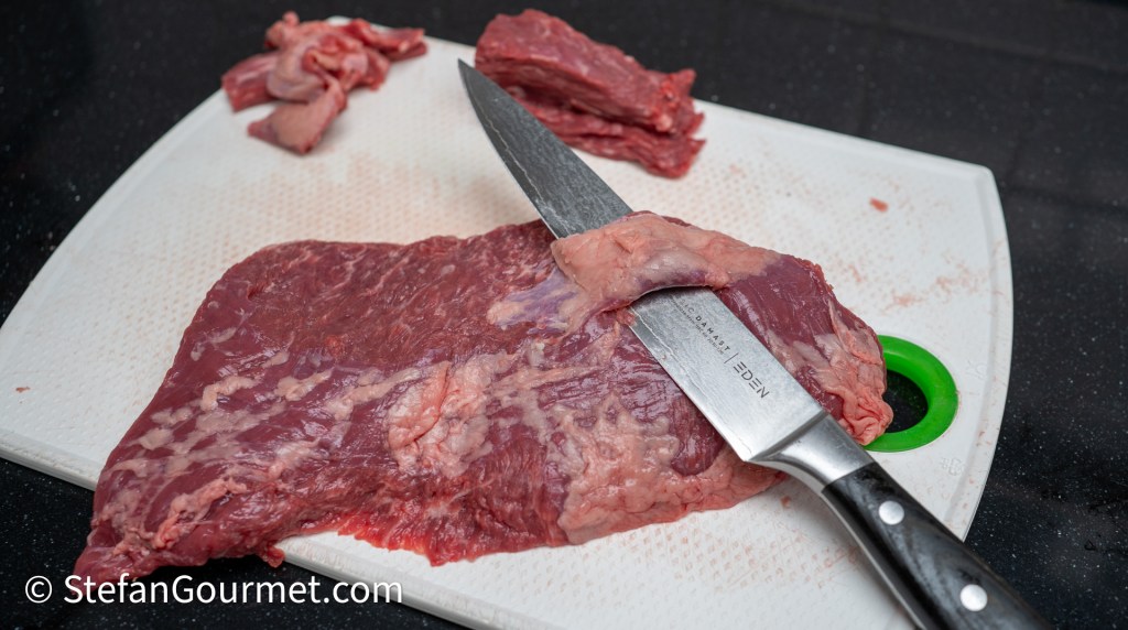Raw meat on a cutting board with a knife, surrounded by scattered smaller pieces of meat.