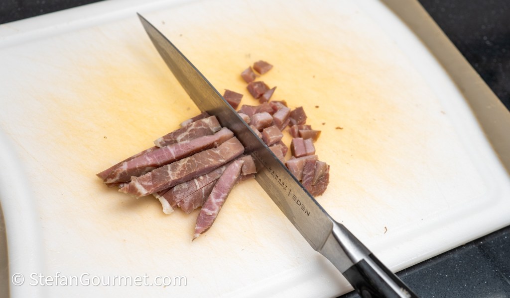 A knife slicing through pieces of ham on a cutting board, with some diced pieces visible.