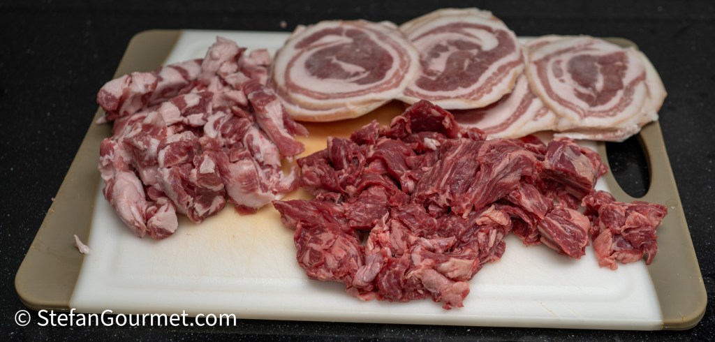 A cutting board with various types of raw meat, including chunks of pork and bacon, arranged neatly.