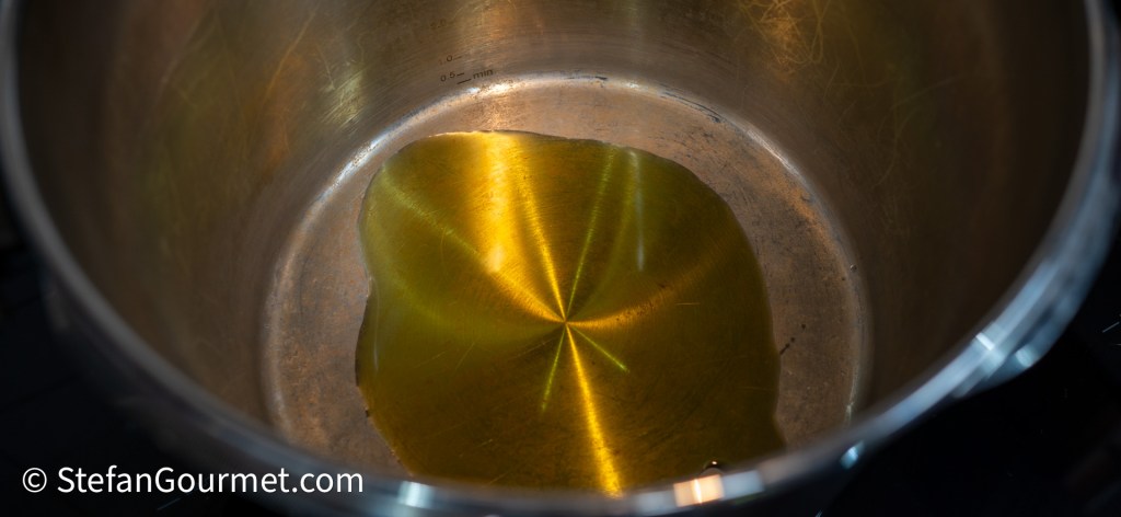 A close-up view of a shiny stainless steel pot with a small amount of golden cooking oil at the bottom.