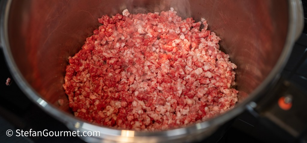 Ground beef being cooked in a stainless steel pot. The meat appears pink and crumbly, indicating it's in the process of browning.