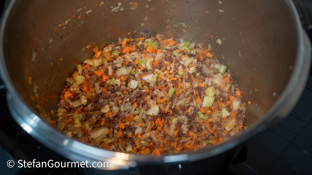 A pot filled with sautéed ground meat and finely chopped vegetables, including carrots, celery, and onions, showing a rich mixture of colors and textures.