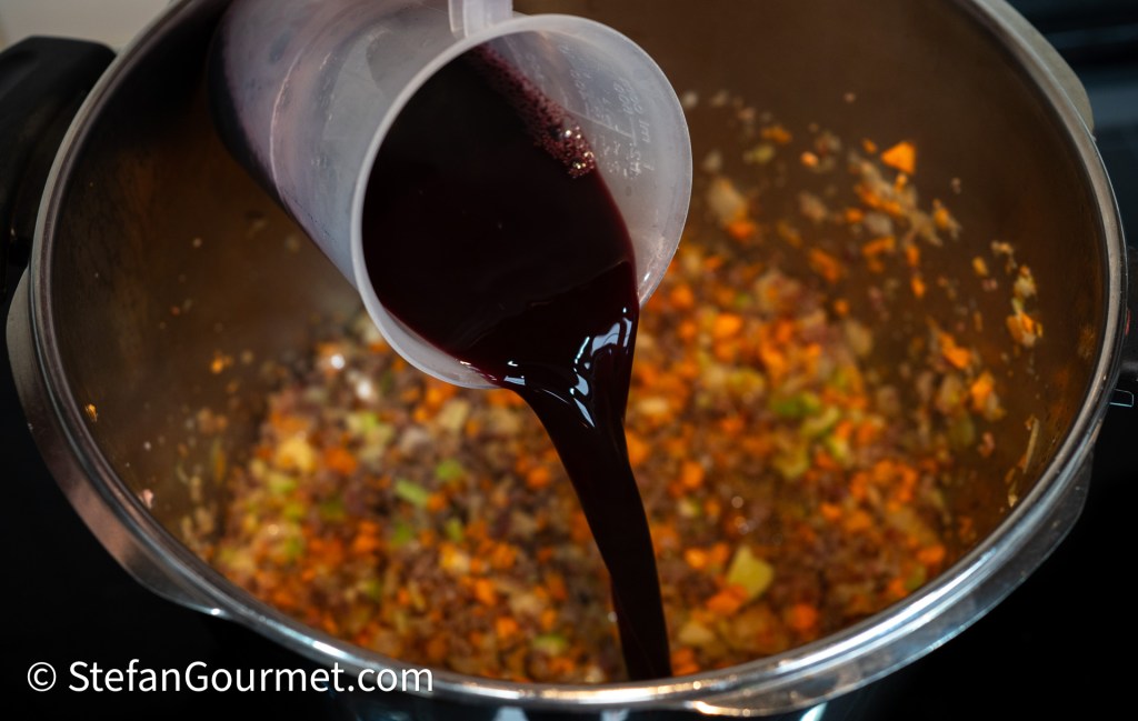 A measuring cup pouring dark liquid into a pot with sautéed vegetables and meat.