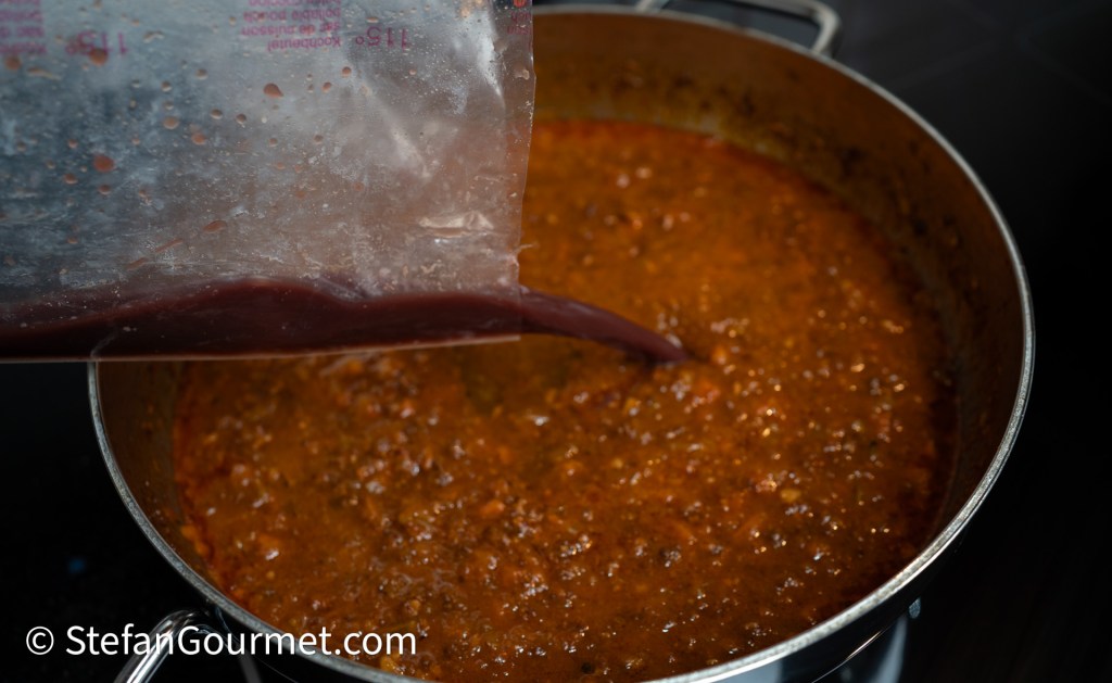 A cook pouring a thick sauce from a plastic bag into a large pot of simmering chili.