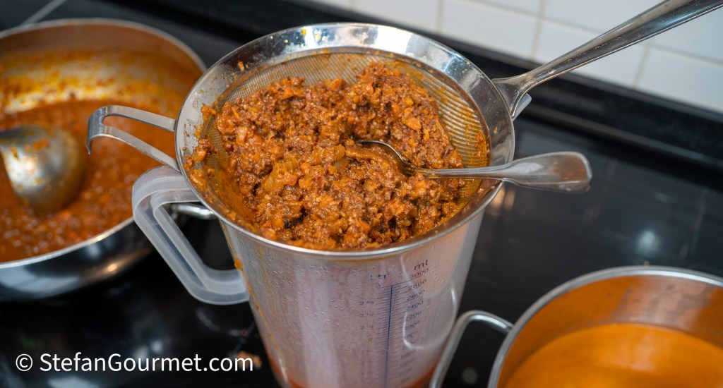 A measuring jug filled with strained meat and sauce mixture, with a spoon resting on the edge, next to a pot of sauce on the stove.