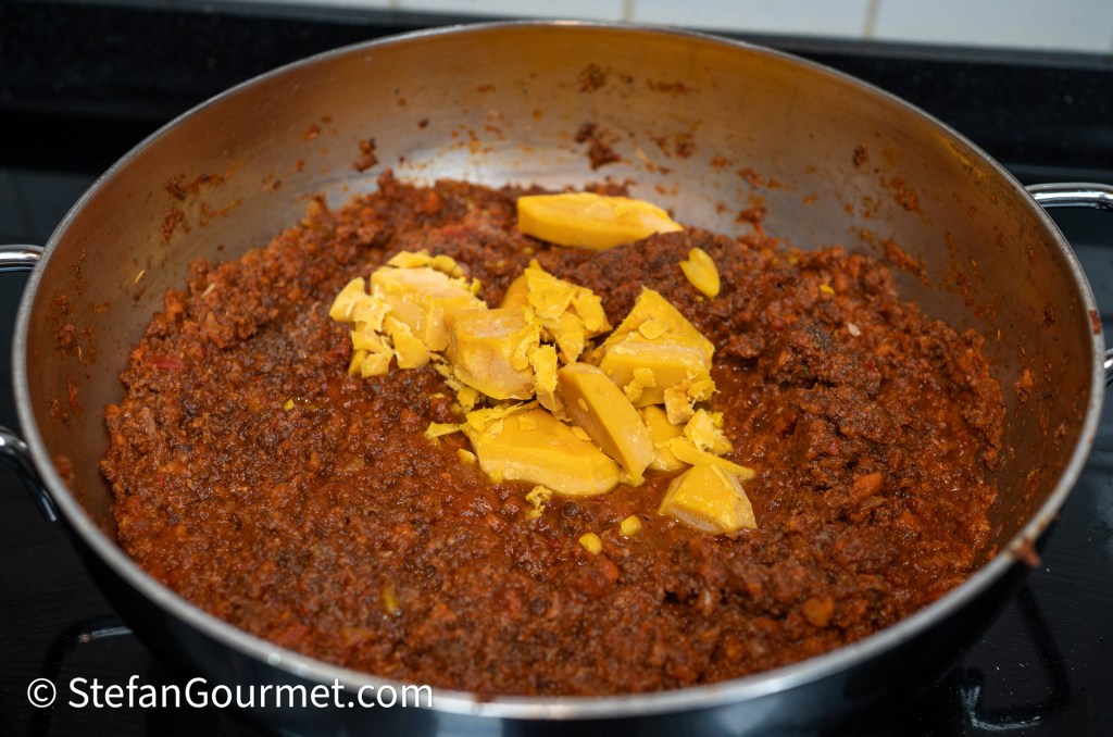 A metallic pan containing a thick, textured mixture of ground meat and spices, with chunks of yellow butter placed on top.