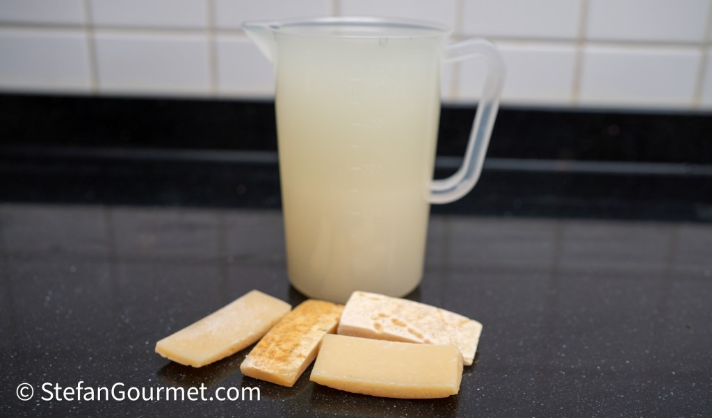 A pitcher of light-colored liquid beside several rectangular pieces of a solid ingredient, likely cheese or a cooking ingredient, on a black countertop.