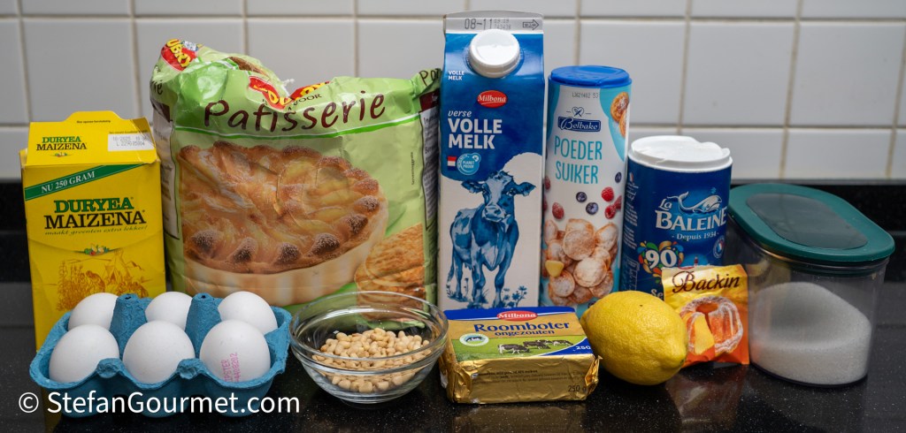 A collection of baking ingredients including eggs, corn starch, whole milk, powdered sugar, butter, a lemon, baking powder, and granulated sugar, arranged on a kitchen counter.