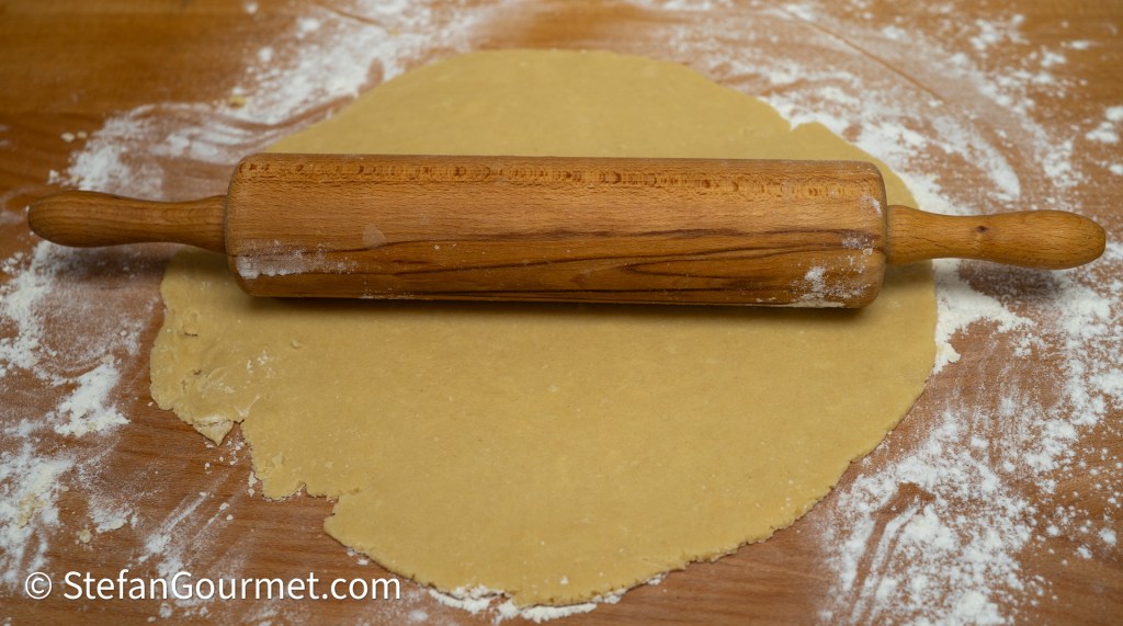 A wooden rolling pin rests on a flat, rolled-out dough on a wooden surface, surrounded by flour.