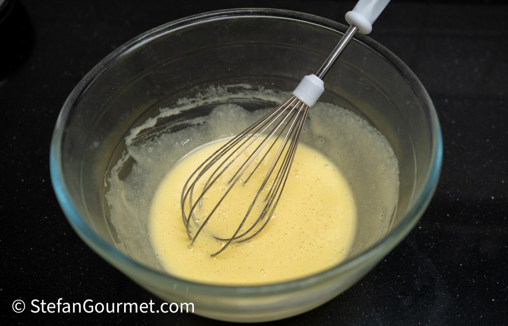 A clear glass mixing bowl with a whisk in it, containing a smooth mixture of beaten eggs on a dark countertop.