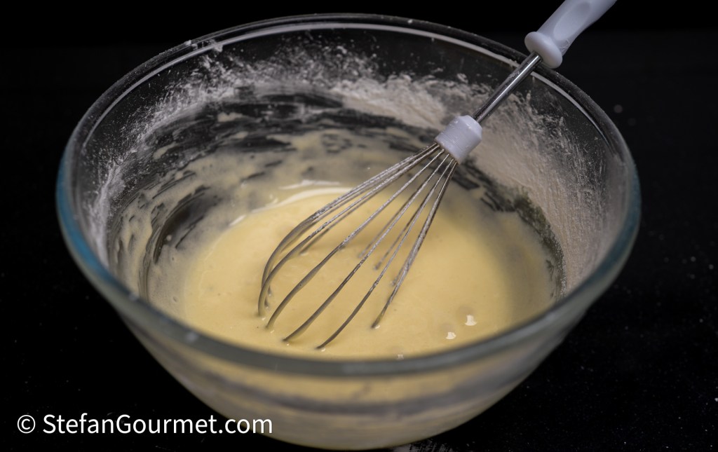A glass bowl containing a light yellow batter with a whisk resting inside, surrounded by flour residue on a black surface.