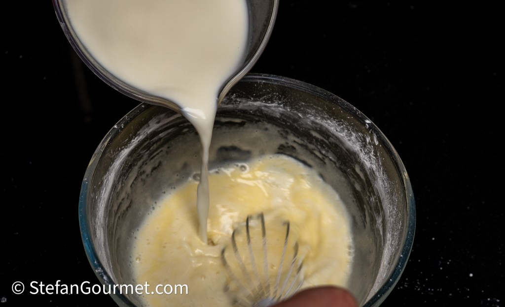 A person pouring liquid into a mixing bowl containing a whisked mixture, with a close focus on the pouring action and the mixed ingredients.