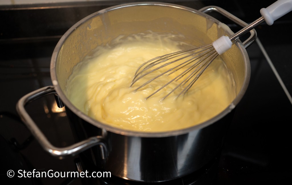 A stainless steel pot on a stovetop with a creamy mixture being whisked inside.