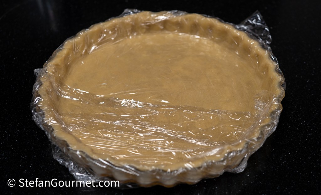 A pie crust covered with plastic wrap on a black surface.