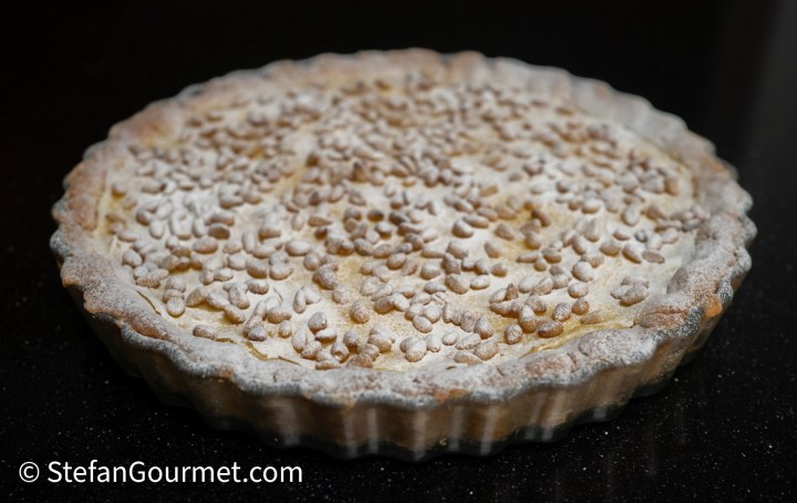 A baked tart with a golden crust, topped with small granules and a dusting of powdered sugar, set against a black background.