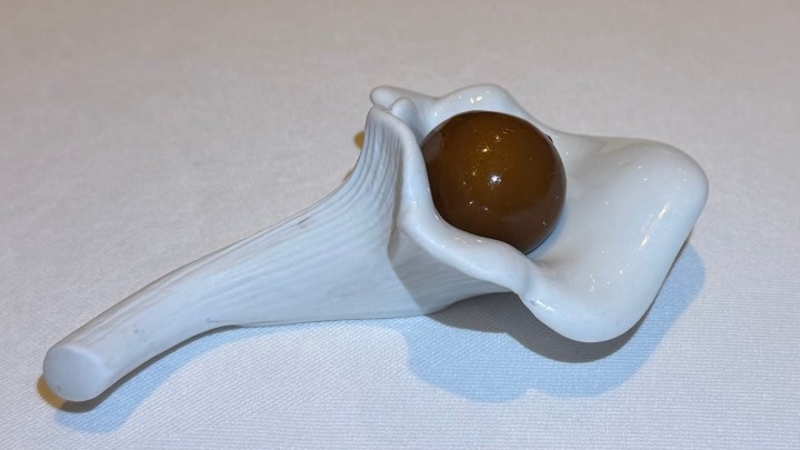 A decorative white porcelain dish shaped like a flower petal holding a round golden-brown sphere.