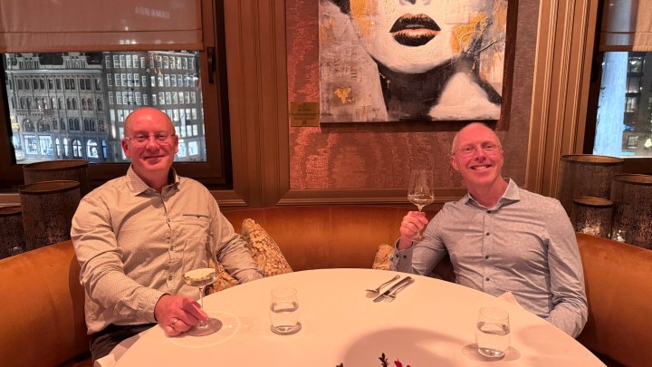 Two men sitting at a round table in a restaurant, each holding a glass of wine and smiling, with an art piece of a woman's face in the background.