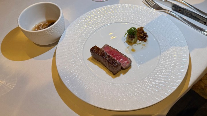 A gourmet dish featuring a perfectly cooked steak, sliced to reveal a pink center, served with a small garnish and a delicate sauce, alongside a small bowl of soup.