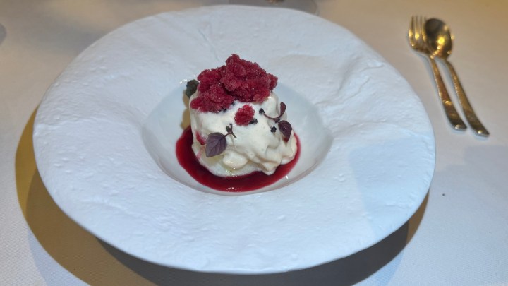 A dessert featuring a scoop of creamy vanilla ice cream topped with raspberry granita and small edible blossoms, served on a textured white plate with a raspberry sauce ring.