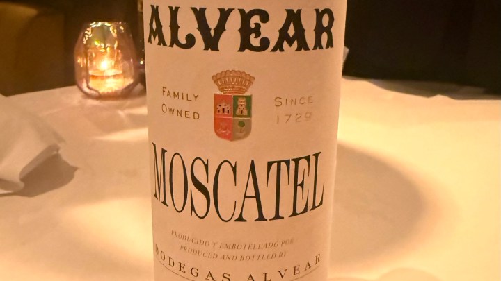 A bottle of Alvear Moscatel wine featuring a white label with black text and colorful crests. The background is softly lit with a candle and blurred restaurant setting.